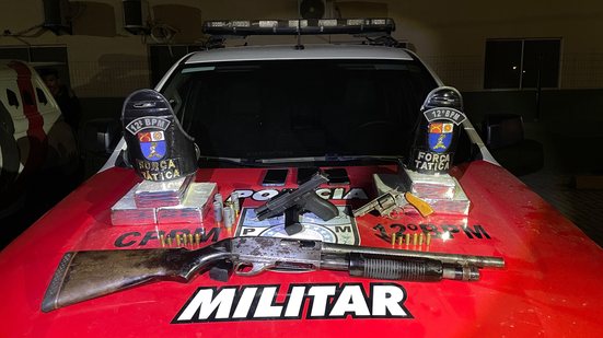 Armas, munições e pacotes de entorpecentes apreendidos expostos sobre o capô de viatura da Polícia Militar.